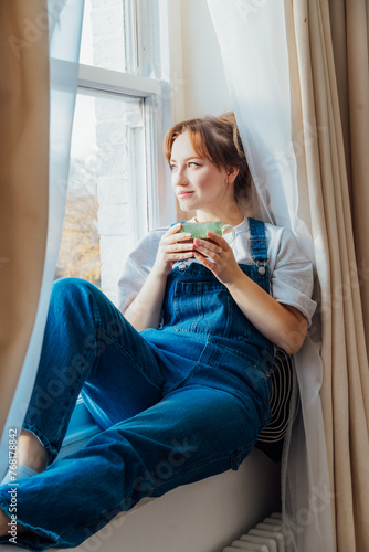 Time To Relax. Young woman looks out the window overlooking the city, sits on windowsill at cozy home holds cup of hot tea drink. Happy calm female taking break for mental health wellbeing. Vertical