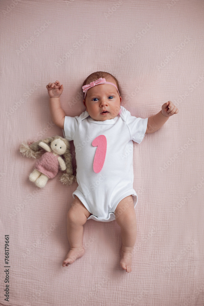 girl baby infant with a bow lies on the bed in a white bodysuit with a doll toy next to it, the child has a pink number 1 months on a pink background
