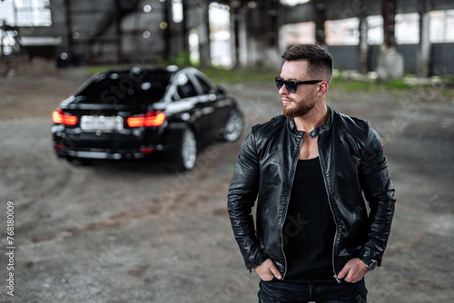 Brutal handsome man in leather jacket with car. Confident young model in stylish sunglasses.