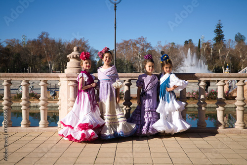 four pretty little girls dancing flamenco dressed in typical gypsy costume pose in a famous square in seville, spain. In the background a large fountain. Flamenco, cultural heritage of humanity. © @skuder_photographer