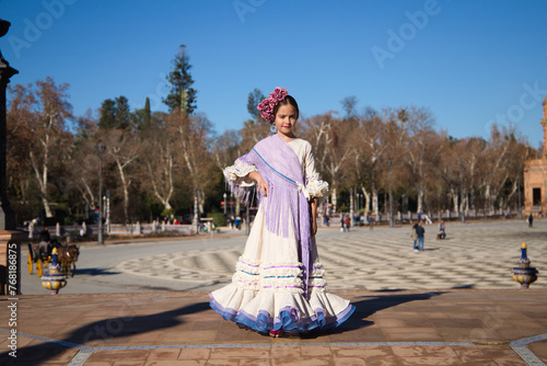 a little girl dancing flamenco dressed in a beige dress with ruffles and purple fringes in a famous square in seville, spain. The girl has flowers on her head and her hair in a bun.