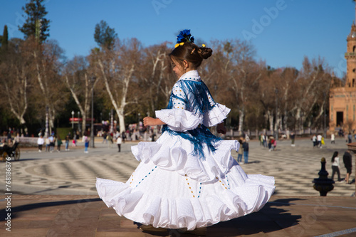 a little girl dancing flamenco dressed in a white dress with ruffles and blue fringes in a famous square in seville, spain. The girl has flowers on her head. Flamenco, cultural heritage of humanity.