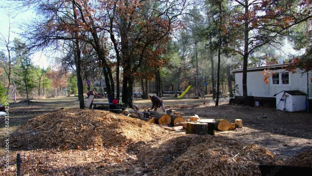 Wide angle shot of pile of mulch and oak logs meanwhile man using ax to process lumber into firewood as smoke rise from manufactured house chimney from burning wood in fireplace. Wide angle shot of pile of mulch and oak logs meanwhile man using ax to process lumber into firewood as smoke rise from manufactured house chimney from burning wood in fireplace.