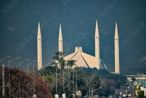 A Faisal mosque with tall minarets, amidst trees and streetlights, against a mountain backdrop under a clear sky. photo