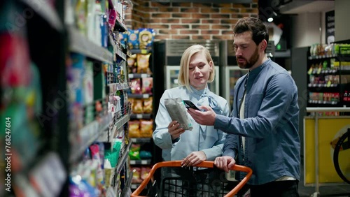 Young caucasian husband standing near shopping cart while attractive wife taking package from shelves in grocery store. Couple dressed in casual attire. Aisle with snacks, colorful bags and boxes.
