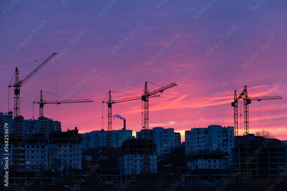 Urban expansion, city expansion. Multiple cranes on building multiple residential houses. Very large construction site at the background of an atmospheric red and blue sky.