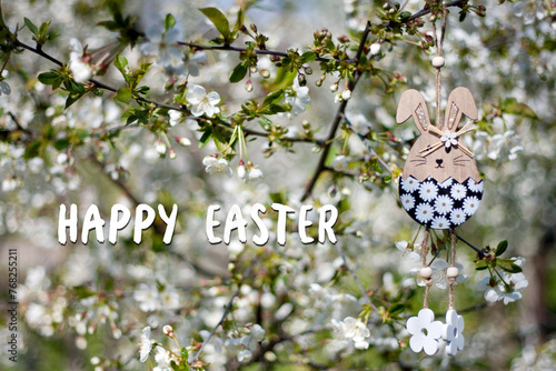 Greeting card with text Happy Easter in English. Easter decor. Decorative wooden Easter bunny toy on background of blooming spring garden. Outdoor