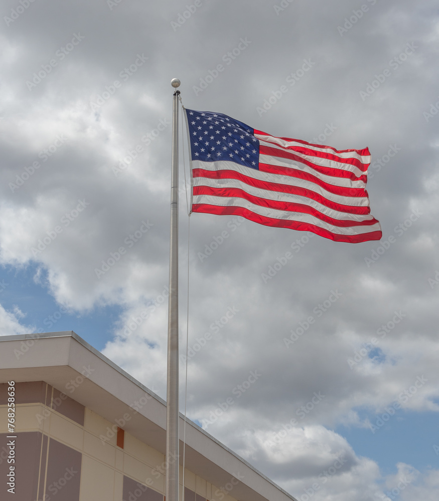American flag waving in wind in front of building with cloudy sky in background