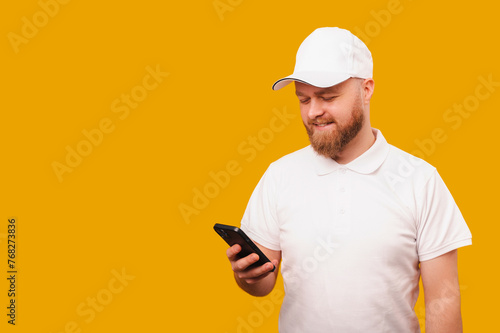 Young smiling bearded delivery man wearing white using phone over yellow background.
