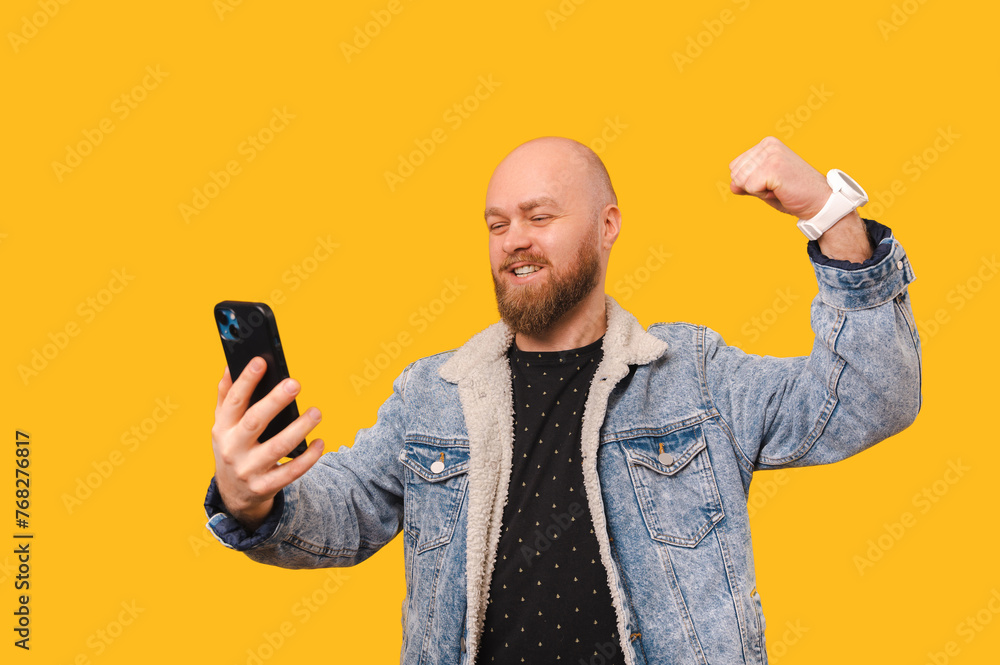 Ecstatic bald bearded man making the winner gesture while looking at his phone over yellow background. Ecstatic bald bearded man making the winner gesture while looking at his phone over yellow background.