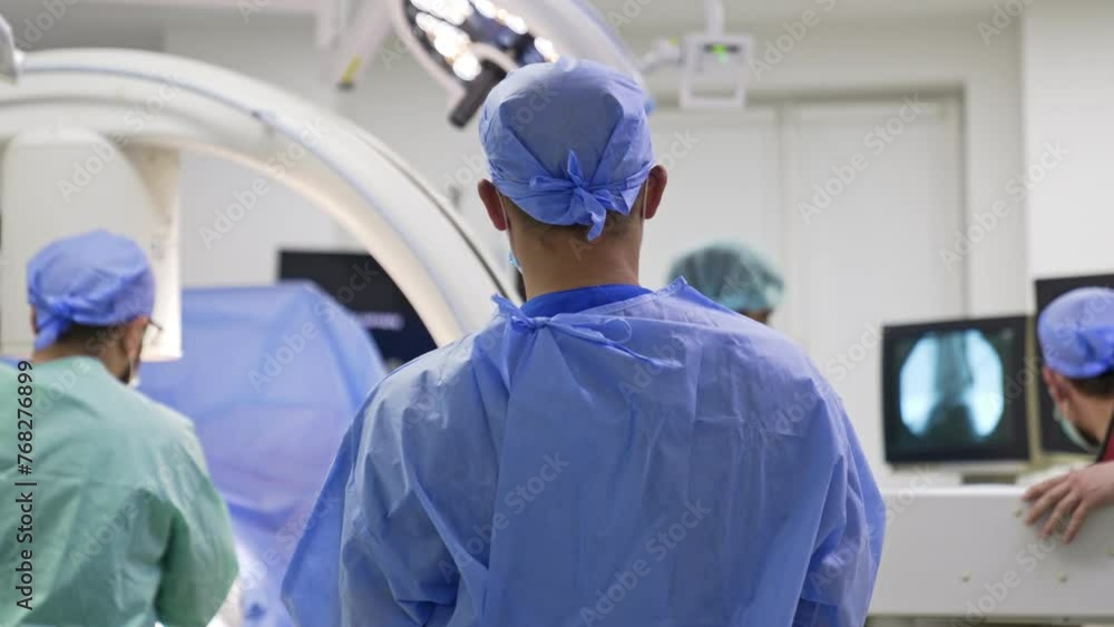 Rear view of a male doctor in blue uniform and cap standing in the surgery room. Medics at backdrop check the x-ray scans of a patient. Rear view of a male doctor in blue uniform and cap standing in the surgery room. Medics at backdrop check the x-ray scans of a patient.