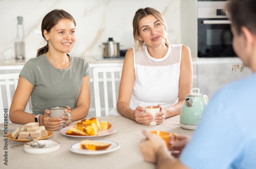 Smiling hospitable woman and young girl receiving male guest over cup of tea with sweets at home, sitting at cozy kitchen table and talking friendly..