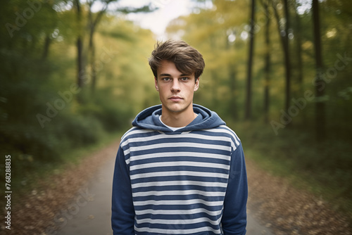 Serious young man in striped hoodie standing on forest path, introspective autumn mood, youth in nature. © Darya