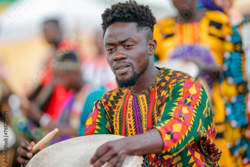 An African man in colorful attire focuses intently as he plays a traditional drum, emitting a sense of rhythm and culture