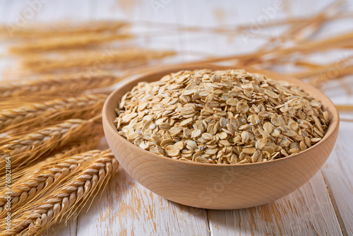 Raw dry rolled oats are scattered out of the wooden bowl on a light table, selective focus.