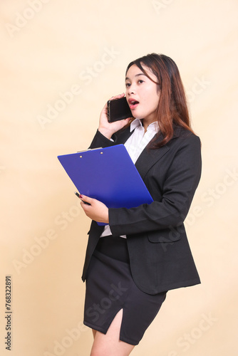 Beautiful formal indonesia executive woman in suit standing surprised and pose hugging clipboard and telephone with smartphone gadget candid on yellow background for commercial, vacancy and business