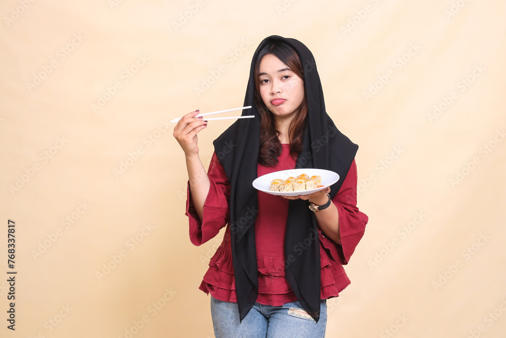 Beautiful mature indonesia woman in red dress wearing hijab disappointed with holding chopsticks to the side and carrying a plate containing dimsum (Chinese food). used for food and lifestyle content Beautiful mature indonesia woman in red dress wearing hijab disappointed with holding chopsticks to the side and carrying a plate containing dimsum (Chinese food). used for food and lifestyle content