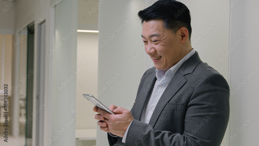 Asian middle-aged smiling businessman chinese japanese korean happy business man employer CEO executive manager near wall in office hall browsing mobile phone scrolling smartphone chatting online app Asian middle-aged smiling businessman chinese japanese korean happy business man employer CEO executive manager near wall in office hall browsing mobile phone scrolling smartphone chatting online app