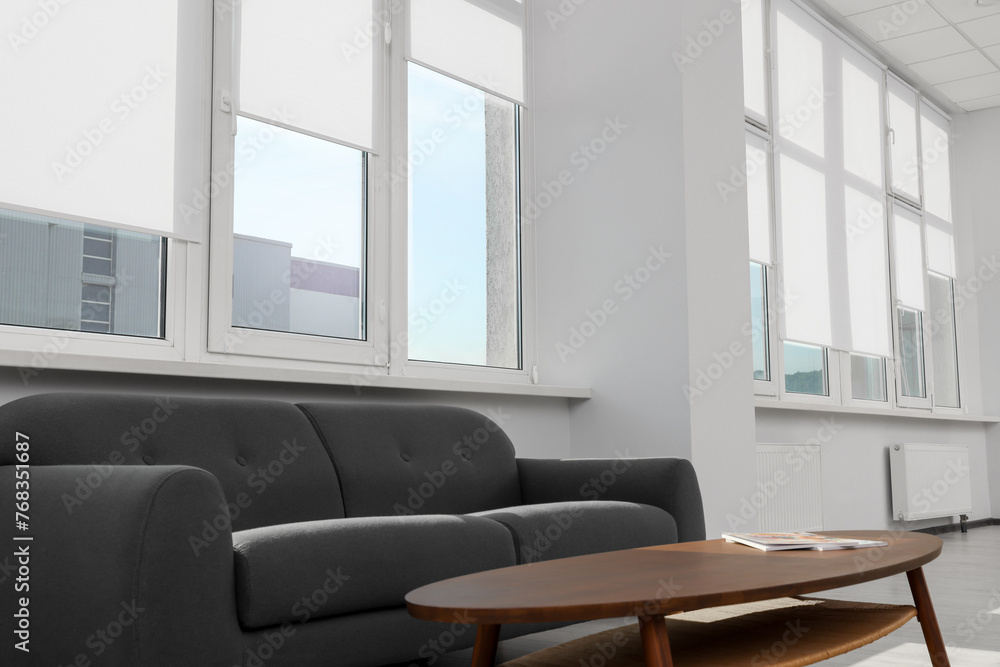Fototapeta premium Comfortable sofa and table near large windows with white roller blinds indoors