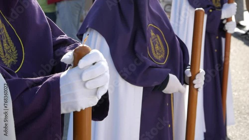 Closeup of three penitent in line formation dressed on traditional white and purple tunics ready to march. They hold long candles with gloves. Camera tilts down showing their robe's. Holy week.