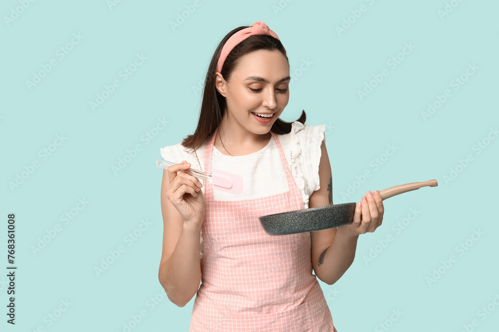 Portrait of happy young housewife in apron with frying pan and spatula on blue background Portrait of happy young housewife in apron with frying pan and spatula on blue background