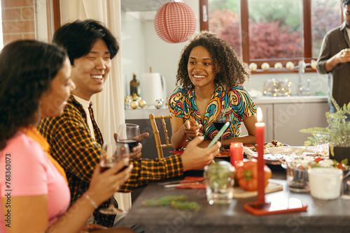 Group of friends sitting around festive dinner table at home, eating and use mobile phone