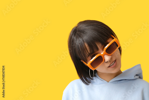 Stylish young woman in sunglasses with nose piercing on yellow background