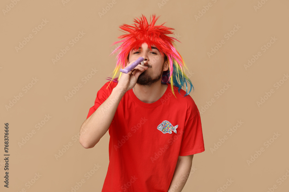 Handsome young man in funny disguise with party whistle and paper fish on brown background. April fool's day celebration Handsome young man in funny disguise with party whistle and paper fish on brown background. April fool's day celebration