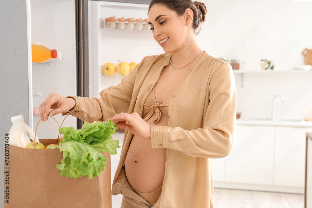 Young pregnant woman with shopping bag near open fridge in kitchen Young pregnant woman with shopping bag near open fridge in kitchen