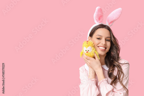 Happy young woman in Easter bunny ears headband with toy chicken on pink background