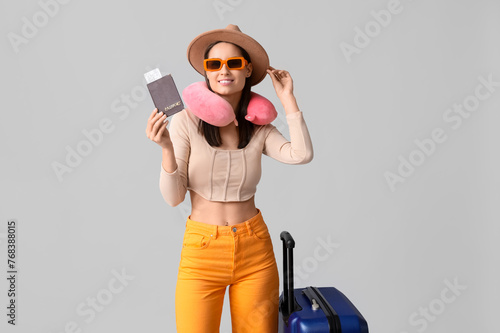 Young happy woman in hat and sunglasses with passport and suitcase on grey background
