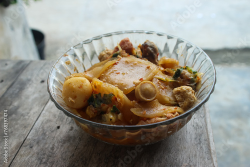 Seblak is a typical Indonesian dish with a savory and spicy taste. cooked with vegetables, sausage, eggs, chicken feet, seafood and processed beef. a bowl of seblak on a wooden table. photo