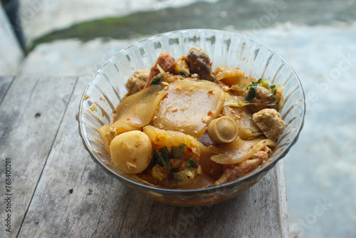 Seblak is a typical Indonesian dish with a savory and spicy taste. cooked with vegetables  sausage  eggs  chicken feet  seafood and processed beef. a bowl of seblak on a wooden table.