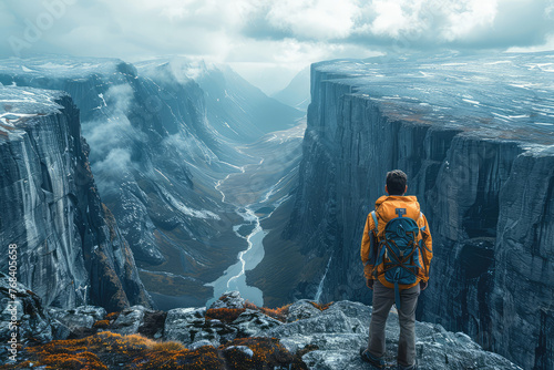 A man hiking stands on the edge of an endless mountain canyon  looking out over rugged terrain and icy waters below. The sky is cloudy with hints of stormy weather. Created with Ai