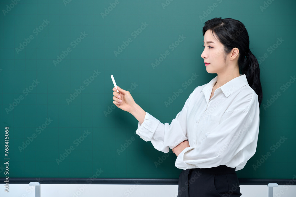 Wearing white heating, a beautiful Asian female teacher is holding a chalk in front of a green chalkboard and giving an Internet lecture with a confident expression and pose. Wearing white heating, a beautiful Asian female teacher is holding a chalk in front of a green chalkboard and giving an Internet lecture with a confident expression and pose.