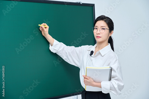 A beautiful Asian female teacher in white heating and glasses is holding a chalk in front of a green chalkboard and giving an Internet lecture with a confident expression and pose.