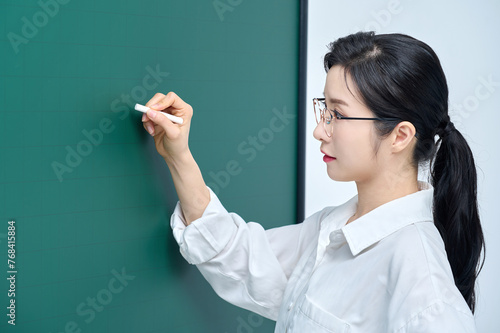 A beautiful Asian female teacher in white heating and glasses is holding a chalk in front of a green chalkboard and giving an Internet lecture with a confident expression and pose.