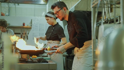 Professional chef holding skillet and using blow torch to cook dish in fire flames when working with female colleagues in restaurant kitchen photo