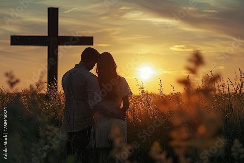 Couple praying God together in field in front of cross at sunset.