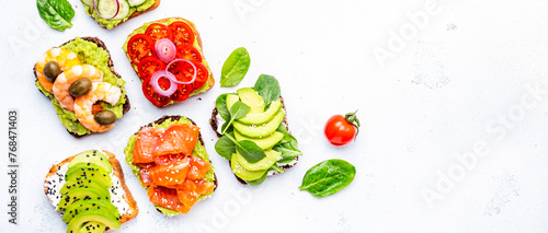 Avocado toasts with salmon, shrimp, vegetables, spinach, capers and cream cheese, white table background, top view