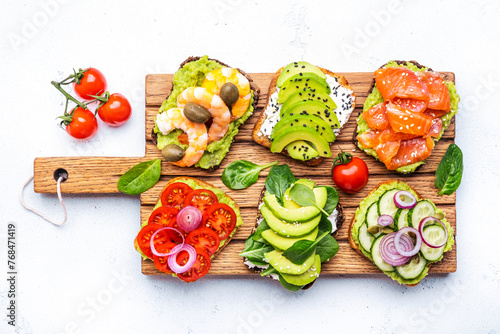 Avocado toasts with salmon, shrimp, vegetables, spinach, capers and cream cheese, served on wooden board, white table background, top view