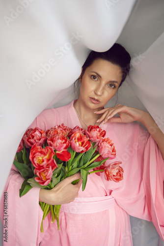 Beautiful portrait brunette woman in a pink Asian dressing gown standing in front of the window against the curtains with red tulips for a holiday.