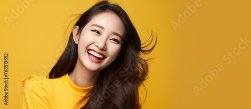 A young Asian woman with long hair is smiling brightly while wearing a yellow shirt. Her expression radiates joy and positivity.