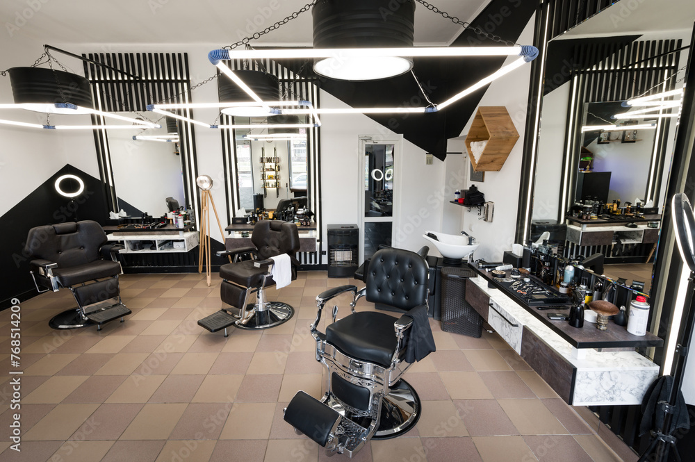 Fototapeta premium The modern interior of a mens barbershop with chairs is shot at a wide angle. Designer interior of a hair salon in black and white colors with LED lamps.