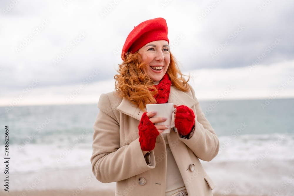 Obraz premium Woman beach in red beret scarf and mitts holding a cup of tea in his hands. Depicting beach relaxation and cozy attire. Walks by the sea