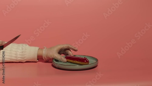 Man hands cut with knife a cherry eclair on the plate. Coral background. Hand turnes a piece of cutted eclair. Cheneese food.
