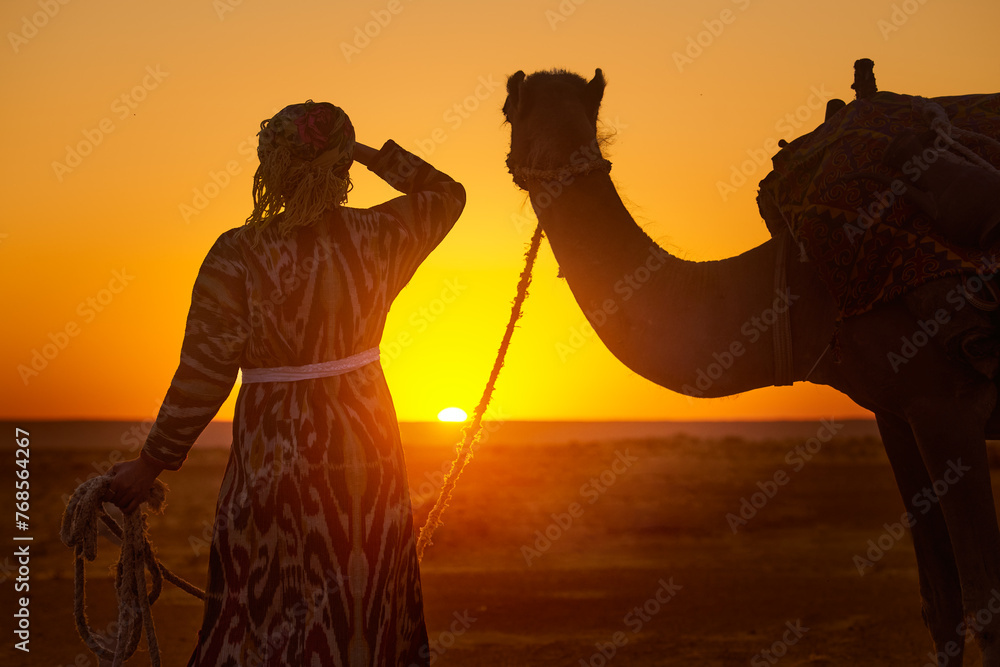 Silhouette of woman in traditional national clothes holding camel by rope while admiring sunset in desert - obrazy, fototapety, plakaty 