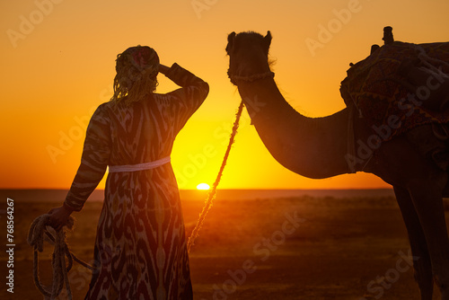 Silhouette of woman in traditional national clothes holding camel by rope while admiring sunset in desert