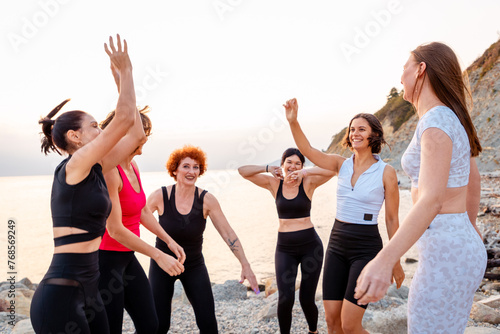 Group of adult athletic multiethnic women in sportswear happily jumps at coast in background of sunset and sea. Concept of wellness and female friendship