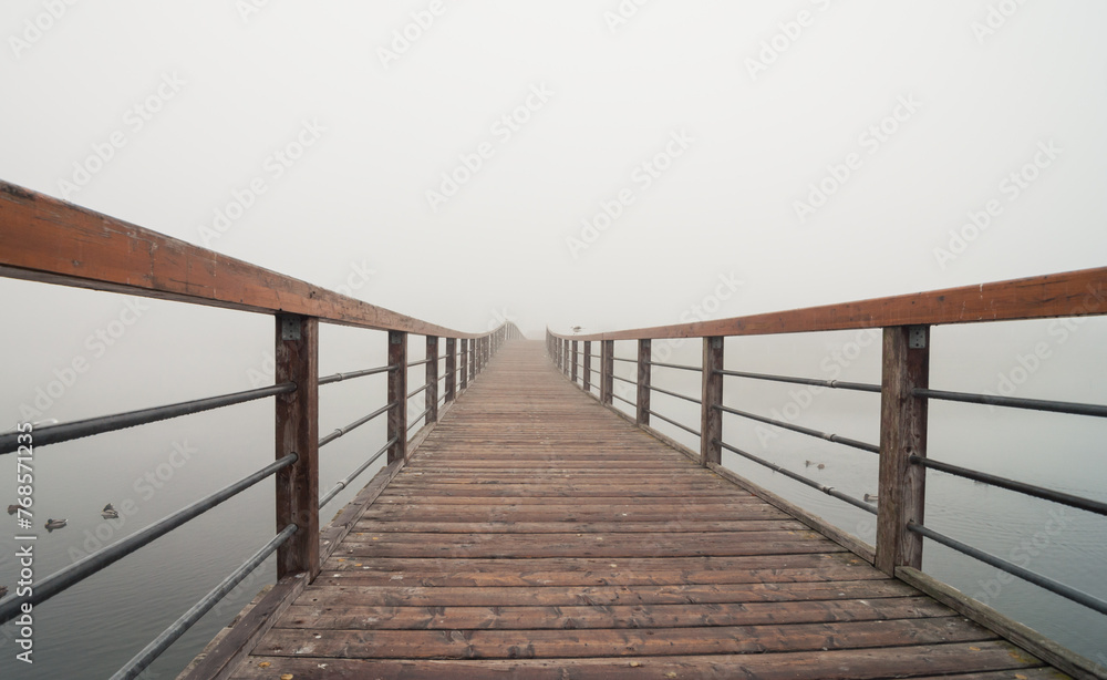 A wooden boardwalk with hand railings over a lake in the fog. Wooden walkway over still water. The walkway curves away from the viewer to the right, disappearing into the fog. Few ducks nearby.